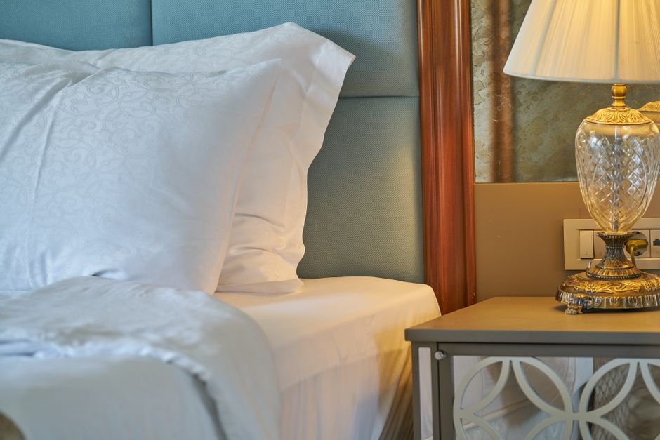 A neatly made hotel bed with crisp white linens, soft natural light streaming through a window, a subtle fold or crease detai